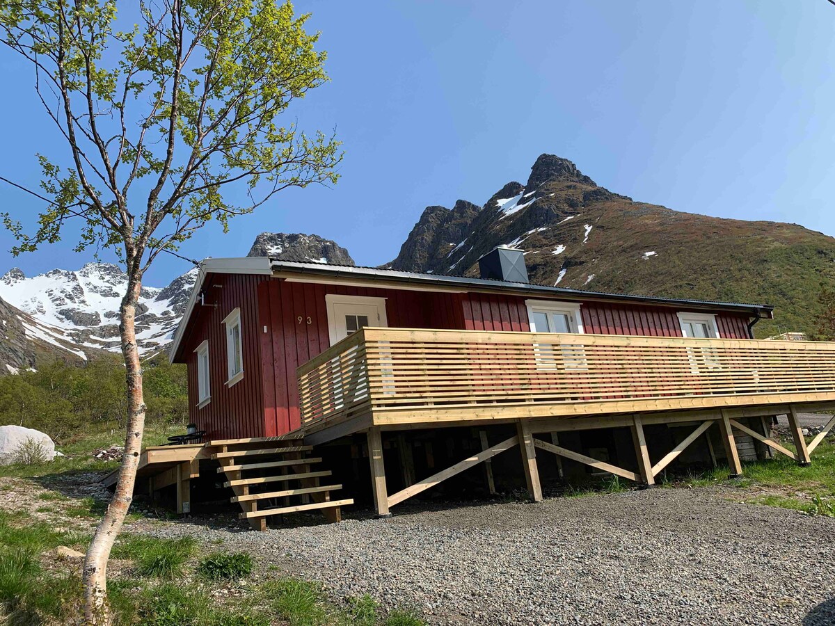 A red cabin is situated on elevated decking, surrounded by mountains. Steps lead to the entrance, and a young tree provides a touch of greenery in the foreground. Snow-capped peaks are visible in the background, hinting at the serene natural environment.