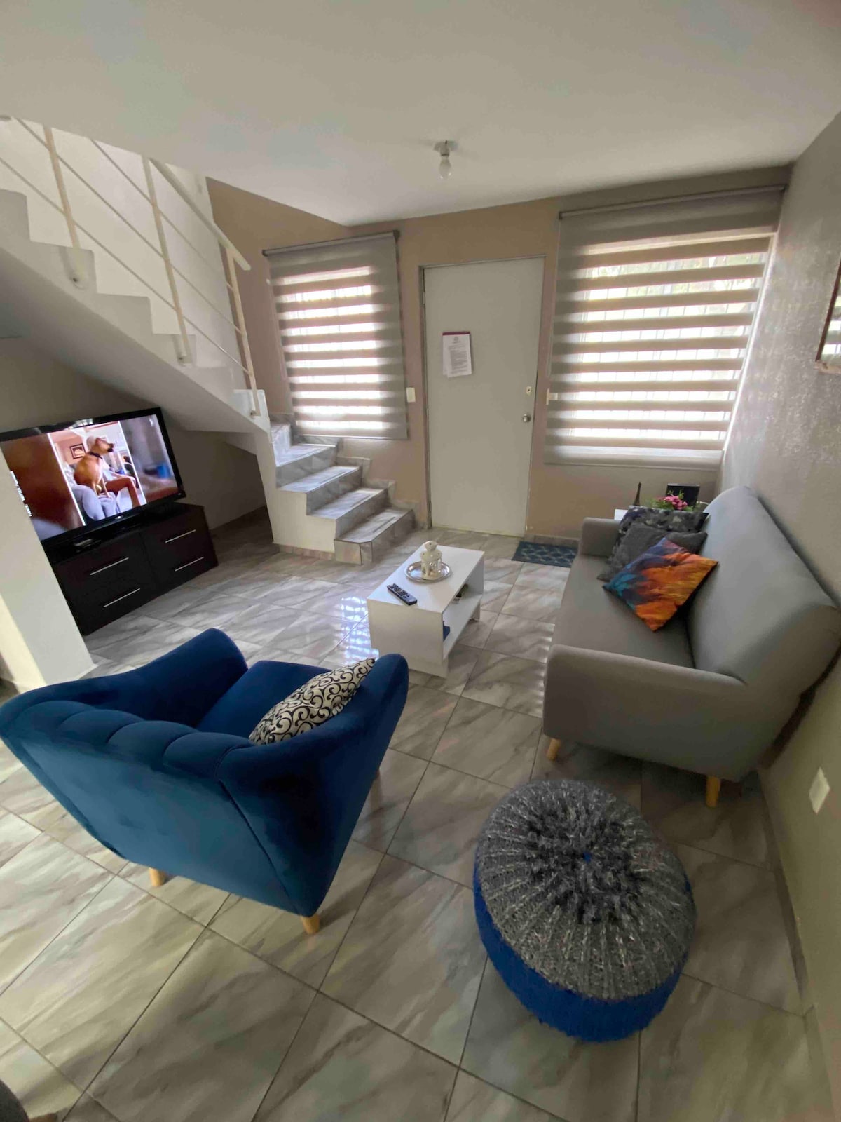 A bright living space is presented, featuring a comfortable gray sofa and a plush blue armchair. A small coffee table sits in the center, surrounded by decorative cushions. A television is positioned on a low cabinet, with light streaming through modern window shades, highlighting the staircase in the background.