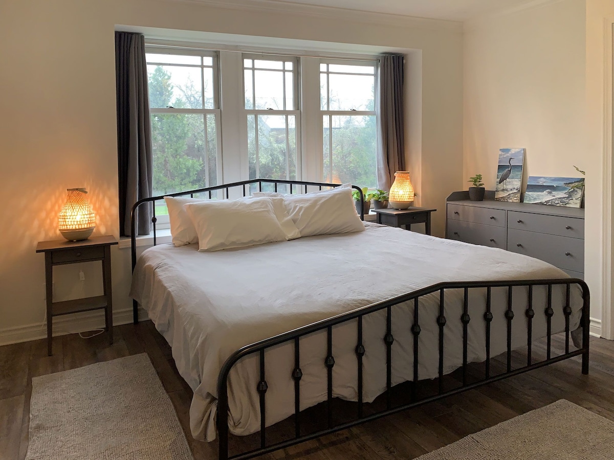 A bright and airy bedroom features a spacious bed with a neatly made white bedding set. Two bedside tables are placed on either side, each with a warm light fixture. Large windows provide ample natural light, framed by dark curtains, and a dresser is positioned against the wall.