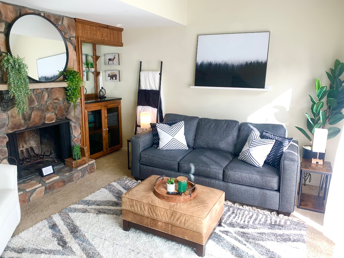 A cozy living area is presented, featuring a dark gray sofa adorned with geometric patterned cushions. A round mirror hangs above a stone fireplace, while a wooden shelf displays framed art. A coffee table with a tray sits on a light area rug, complemented by a tall indoor plant.