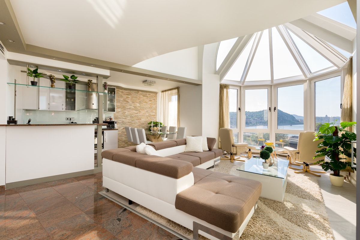 A spacious living area features large panorama windows, providing stunning views of Budapest. A comfortable sectional sofa is positioned around a glass coffee table. Next to the sofa, two high-end armchairs are arranged to enjoy the scenery, complemented by green plants and a modern kitchen area in the background.