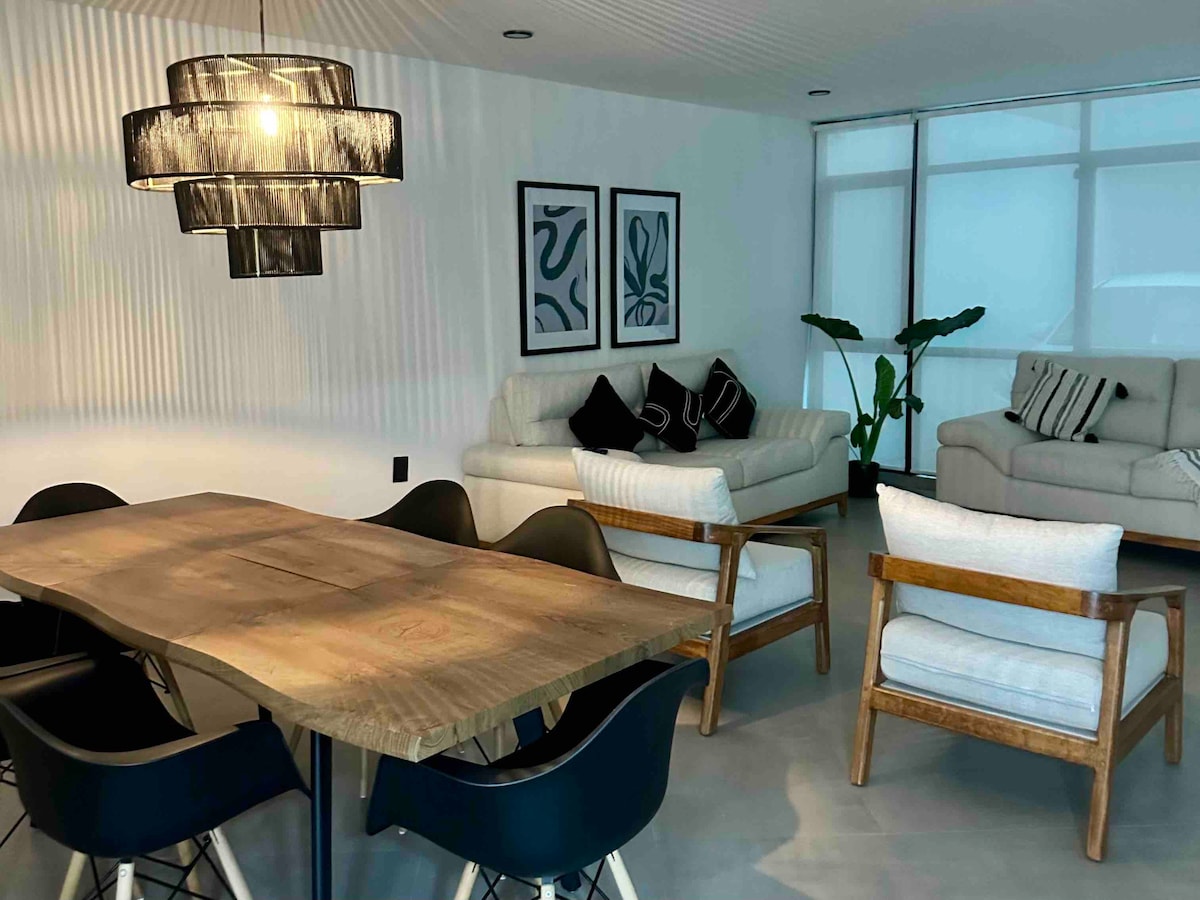 A modern dining area features a large wooden table surrounded by black chairs. Two comfortable armchairs in light wood frame are positioned nearby. A sectional sofa in soft fabric is partially visible, along with black and white wall art accentuating the space.