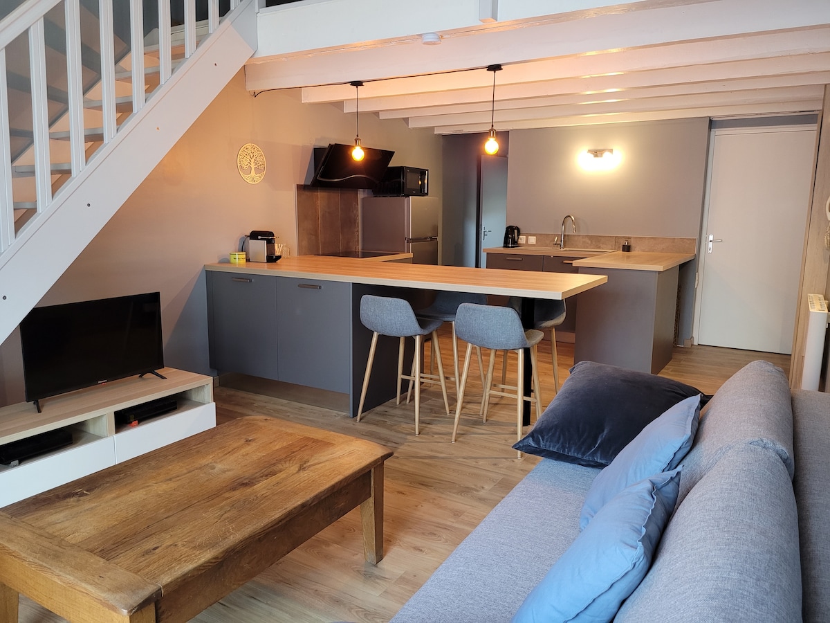 A modern living space is depicted, featuring a cozy sofa and a wooden coffee table. A kitchen area with a large wooden counter and three bar stools is present. A television sits on a white media console, while bright light bulbs hang overhead, illuminating the space.