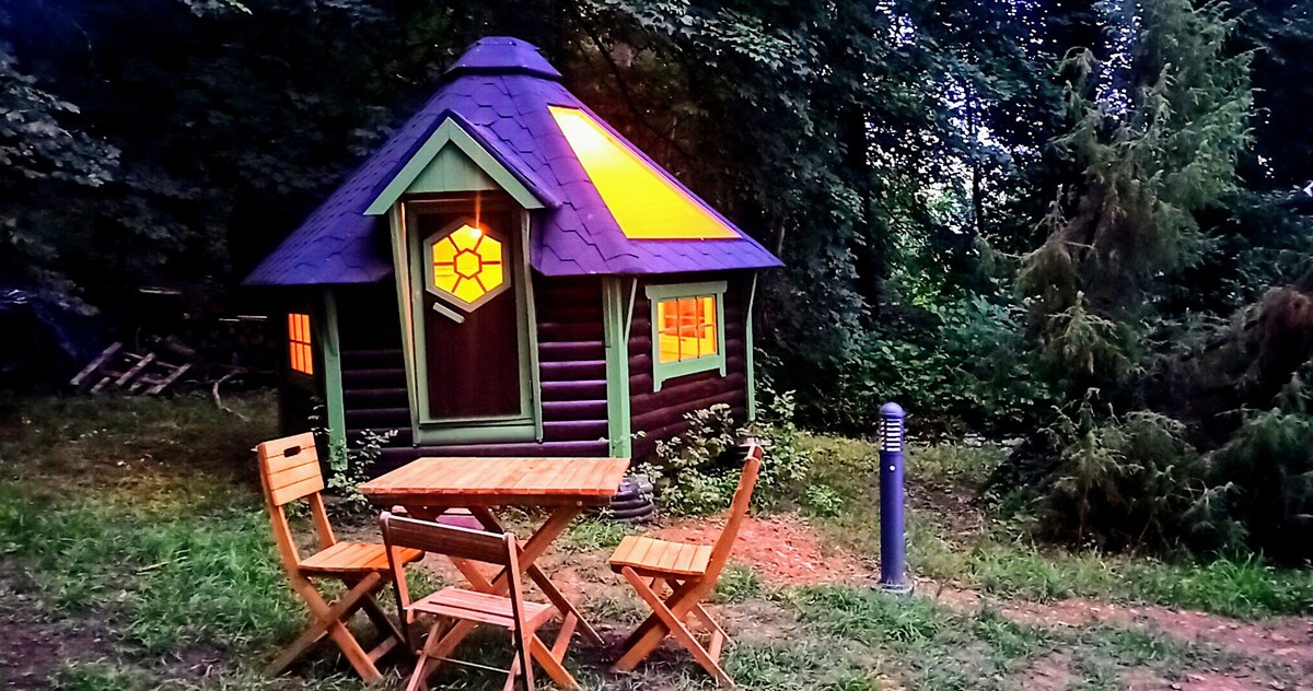 A charming wooden structure is nestled among trees, featuring a unique multi-colored roof and illuminated windows. A small wooden table and two folding chairs are positioned on the grass nearby, offering a cozy spot for outdoor relaxation.