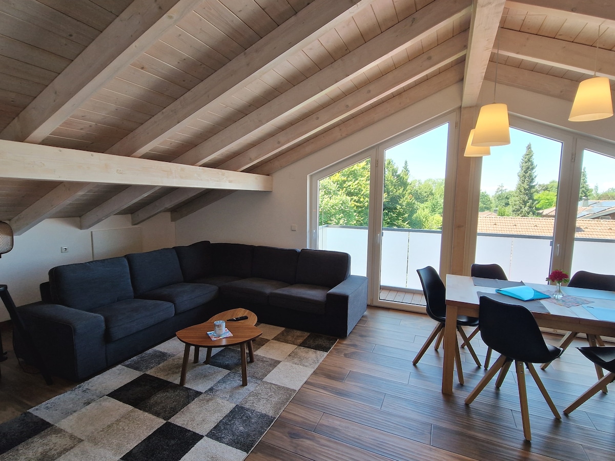 A spacious living area features a large dark sectional sofa and a round wooden table surrounded by four chairs. Large windows allow natural light to fill the space, providing views of the greenery outside. A patterned rug anchors the area, enhancing its inviting atmosphere.