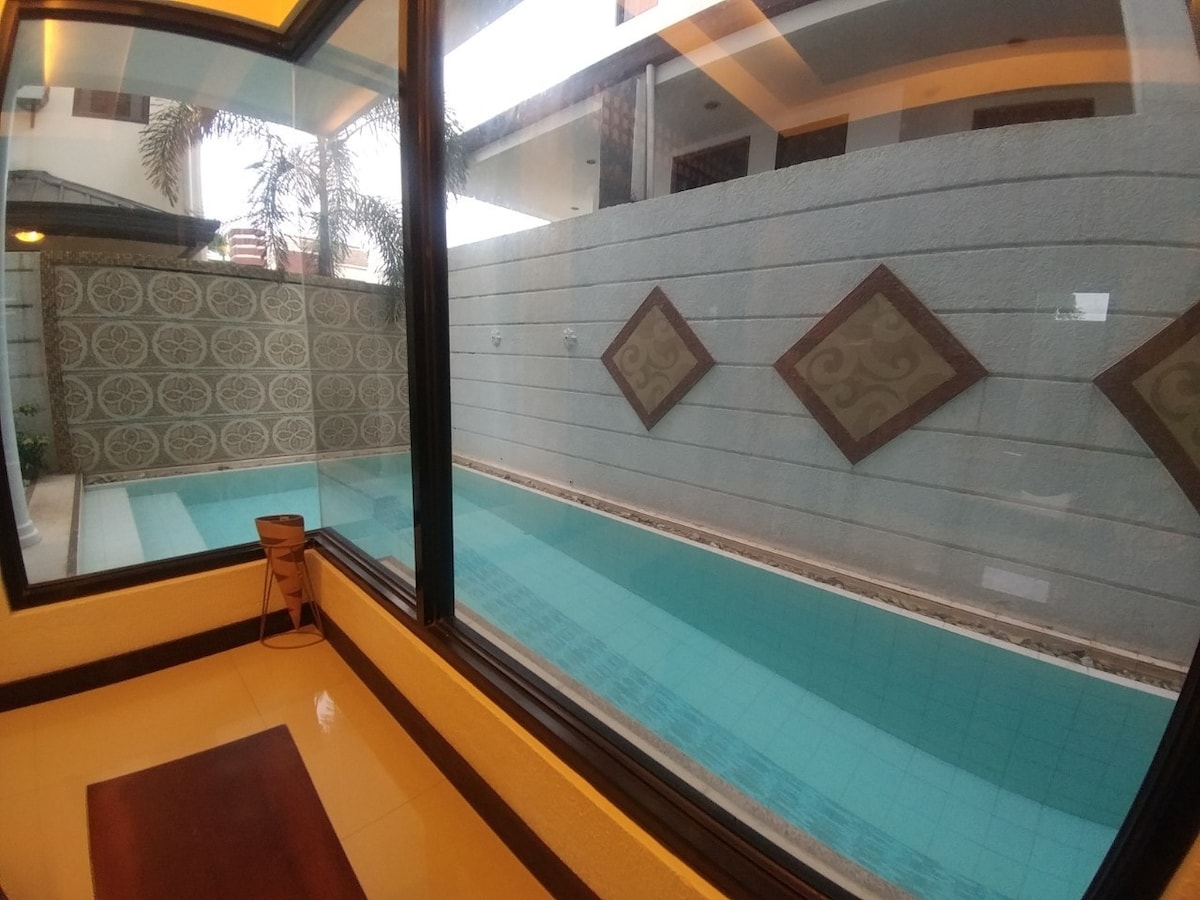 A swimming pool is visible through large glass windows. Its tranquil water reflects the space's soft ambient light. Decorative tiles adorn the pool area, complemented by a minimalist seating arrangement nearby. The view is framed by the surrounding architectural features.