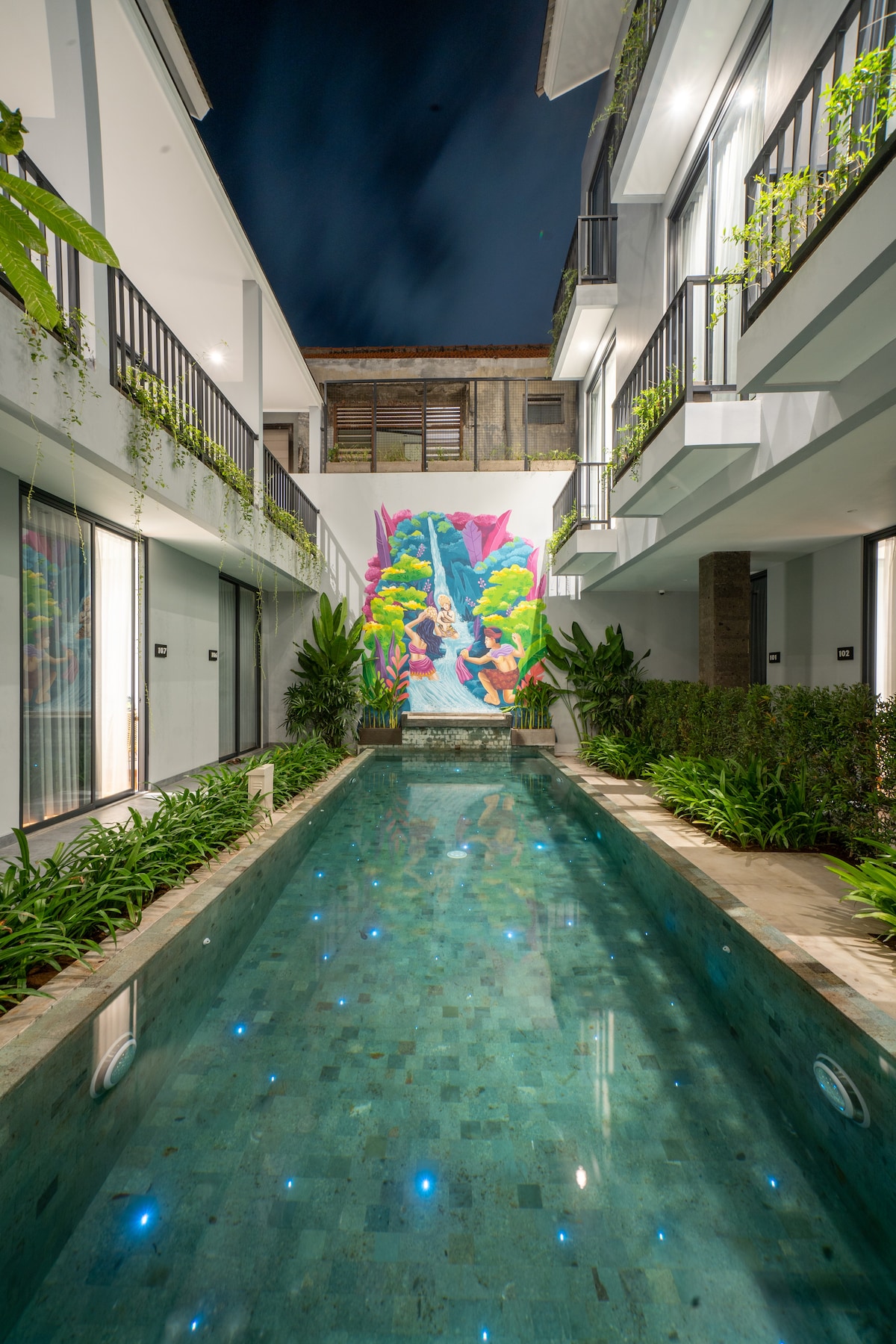 A calm courtyard is presented, featuring a clear swimming pool bordered by lush greenery and decorative plants. Vibrant artwork adorns one wall, providing a colorful focal point. Glass sliding doors lead to the surrounding rooms, creating a bright and open atmosphere.