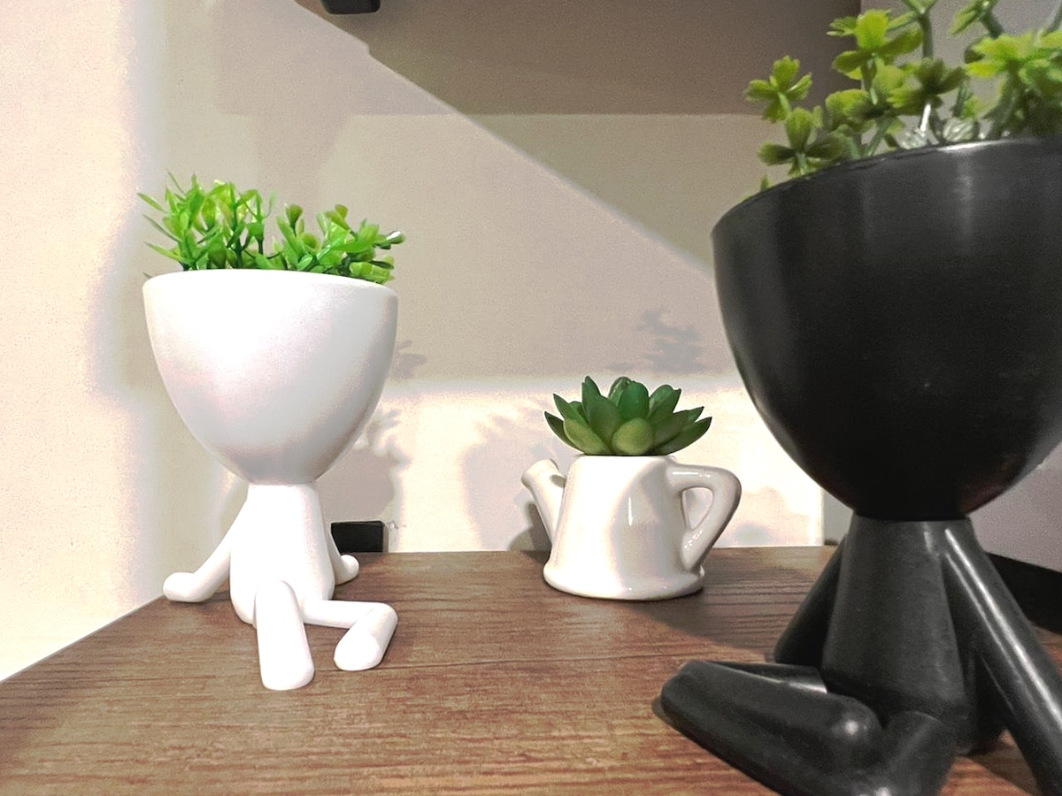 Three unique planters are displayed on a wooden shelf. One planter is white, shaped like a figure sitting, with green plants. A smaller cream-colored planter in the shape of a teapot holds a succulent. A black figure-shaped planter with a round base also features a green plant.
