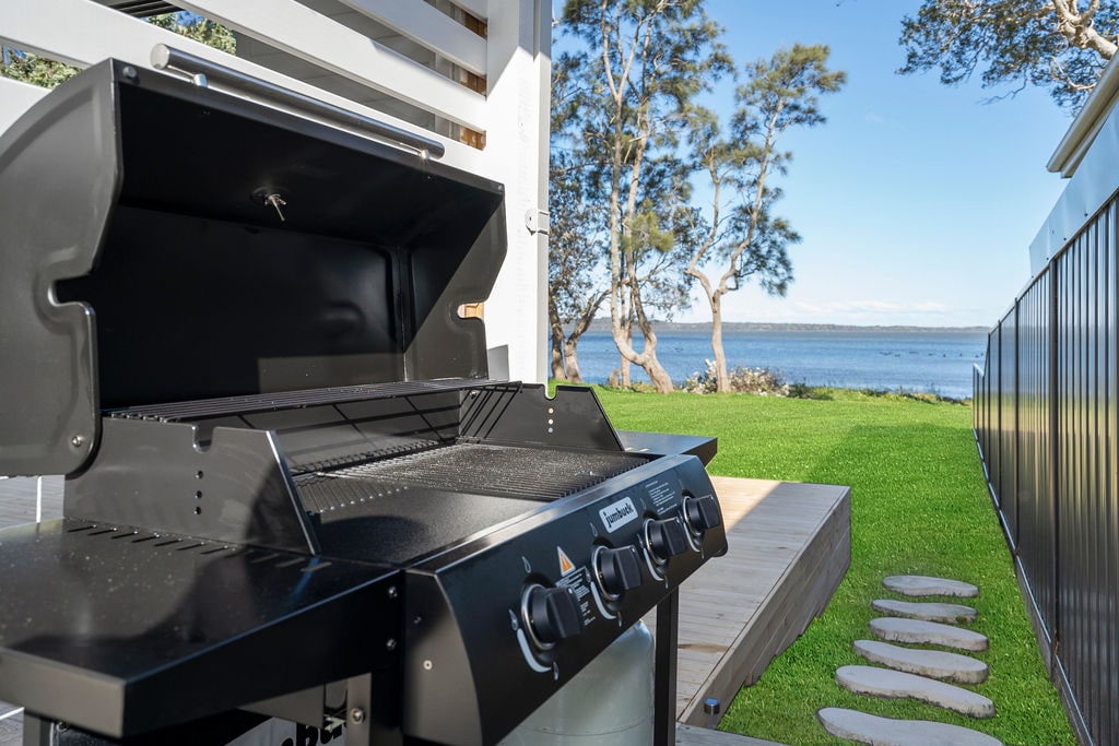 An outdoor barbecue grill is positioned on a patio, facing a serene lake. Soft green grass and stepping stones lead towards the water's edge, framed by gentle trees and a clear blue sky in the background.