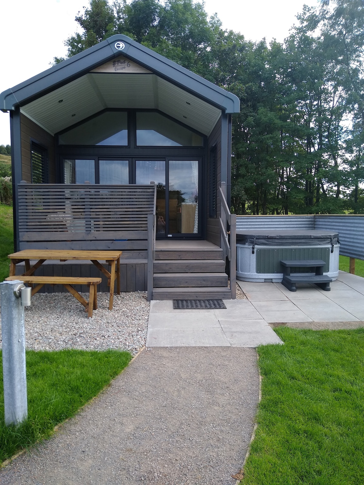A modern retreat is presented, featuring a shaded deck with a picnic table and steps leading to the entrance. An adjacent hot tub is visible beside a paved area, with a path of gravel and grass leading up to the structure. Trees form a natural backdrop.