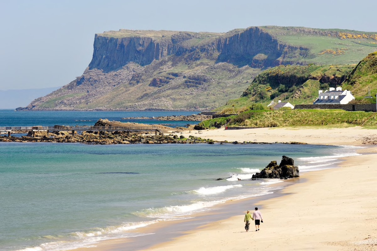 A serene beach scene displays gentle waves lapping against the sandy shore. In the background, a rugged cliffside rises, covered in lush greenery. The coastline is dotted with a few white houses, while a couple is seen strolling along the water's edge.