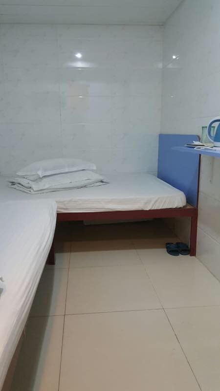 The room features two beds, one a spacious double bed and the other a single bed, positioned separately. Neatly arranged linens are stacked on the double bed. A small shelf displays toiletries, and the floor is tiled in a light color.