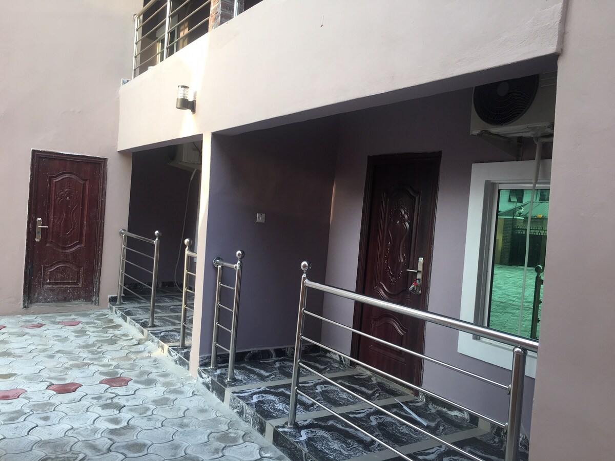 The exterior view showcases a well-maintained walkway that leads to multiple doorways of the apartment units. Each door is framed by modern railings, and natural light illuminates the space. A door to the left has a textured red finish, contrasting with the smooth purple walls.