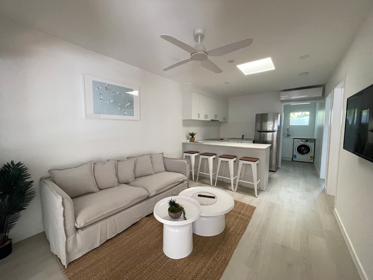 A bright living space is showcased with a neutral color palette. A cozy sofa sits across from a modern kitchenette with stainless steel appliances. Two white side tables are positioned on a textured rug, while a ceiling fan and natural light from the nearby window enhance the ambiance.