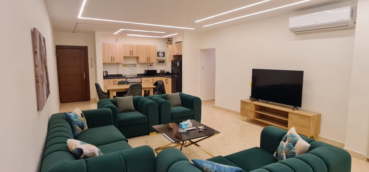 A spacious living area features a generous seating arrangement of green sofas surrounding a central coffee table. A large television is positioned on a wooden stand against the wall. The open kitchen is visible in the background, equipped with modern appliances.