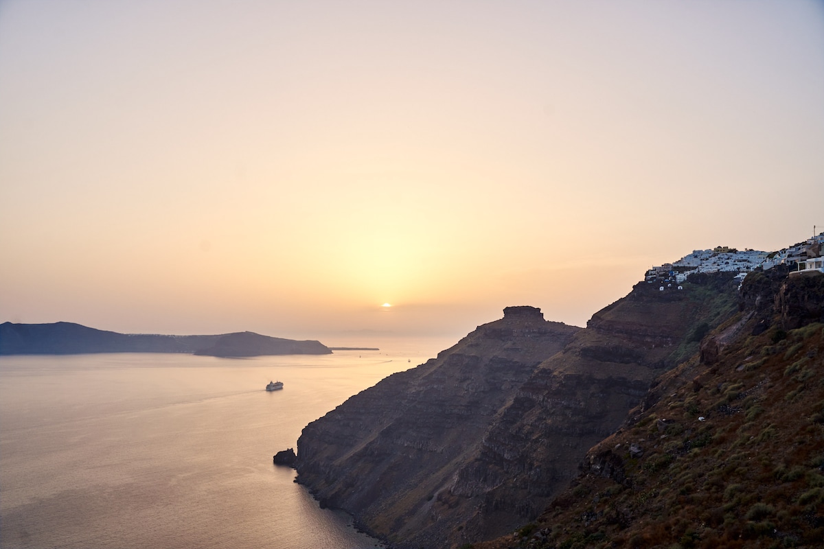 The image captures a tranquil sunset over the Aegean Sea, with soft orange and pink hues illuminating the horizon. The rugged cliffs of Santorini are prominent in the foreground, while a distant boat is seen gently resting on the water's surface.