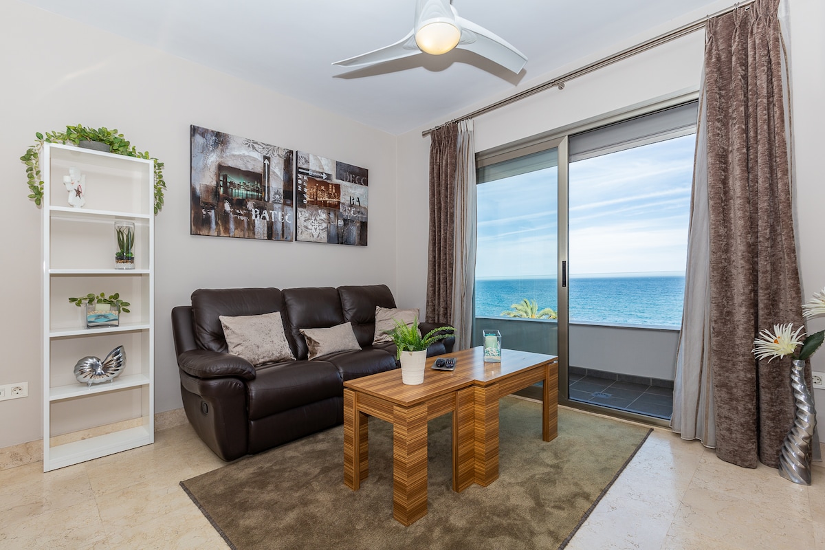 A comfortable living area is seen featuring a dark leather sofa and a wooden coffee table. A large window provides views of the ocean, with light flowing into the space. Decorative plants and a white bookshelf add subtle touches to the modern decor.