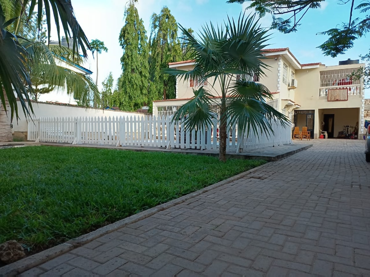 A well-maintained exterior features a spacious lawn bordered by a white picket fence. Multiple palm trees provide shade, while a paved driveway extends towards the villa, revealing a charming two-story building in the background. Natural light enhances the inviting atmosphere.