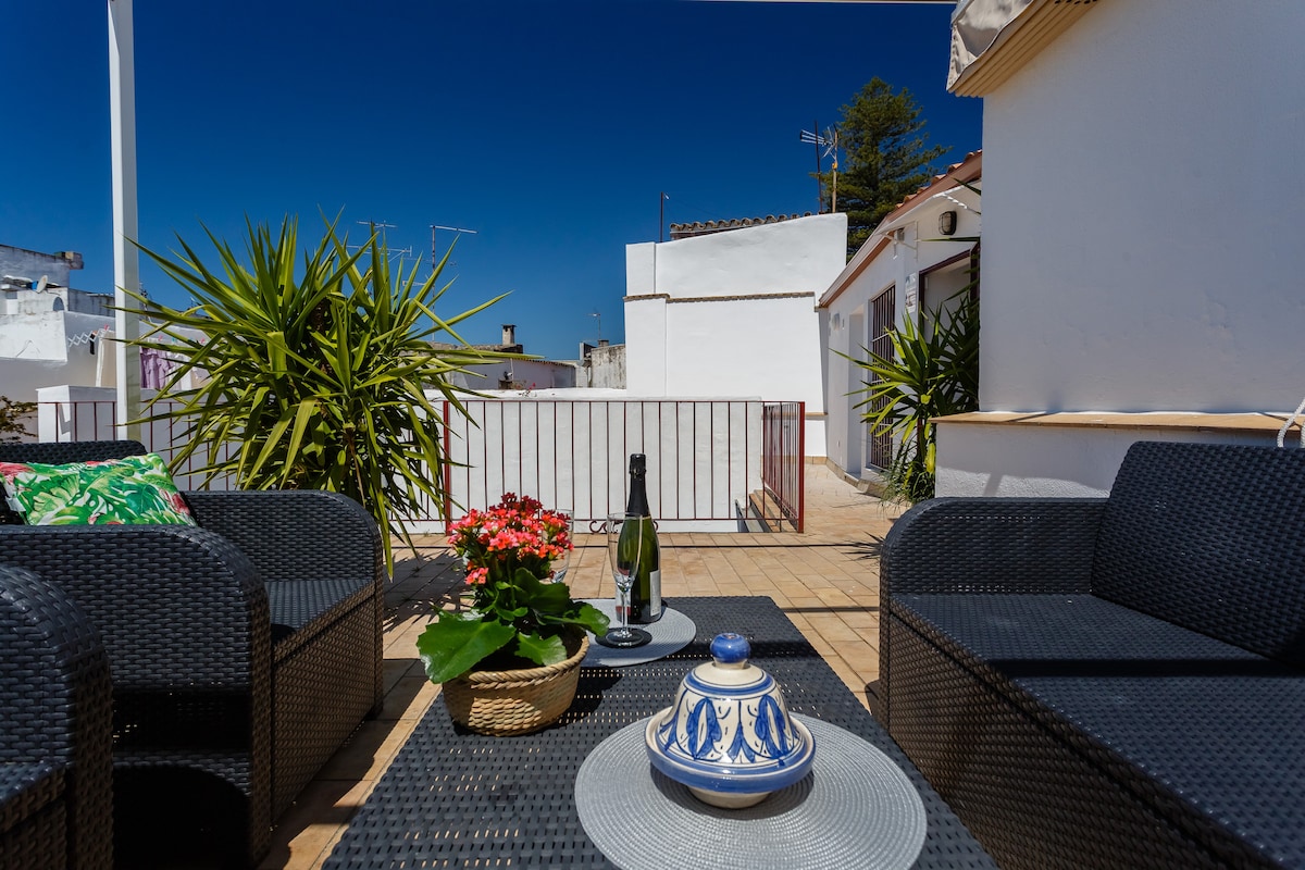 An outdoor terrace is depicted, featuring a seating arrangement with dark wicker chairs and a table. Potted plants add greenery, while a bottle of champagne and a decorative bowl are present on the table. The clear blue sky creates a bright and open atmosphere.