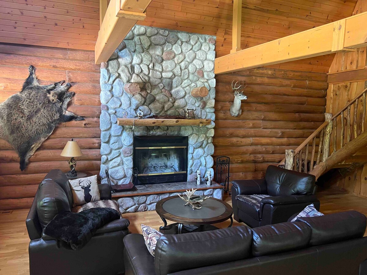The living area features a cozy arrangement of black leather sofas surrounding a circular coffee table. A stone fireplace is complemented by wooden beams, enhancing the rustic feel. Mounted decor includes wildlife elements, adding to the natural ambiance of the handcrafted log home.