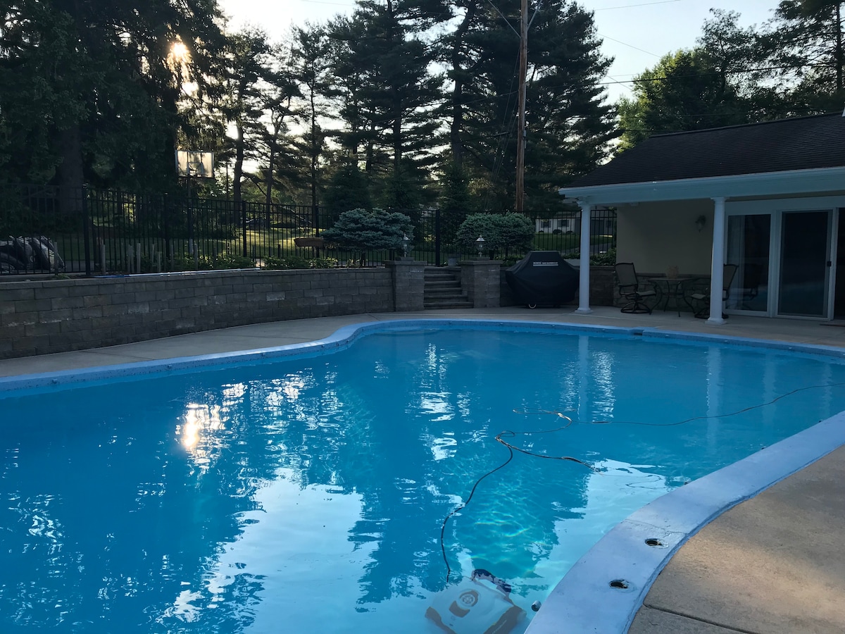 A private, inviting pool area is captured in the image, displaying clear blue water surrounded by a patio and lush greenery. Various seating areas can be seen nearby, along with a barbeque grill positioned against a wall. The serene setting is enhanced by well-maintained landscaping.