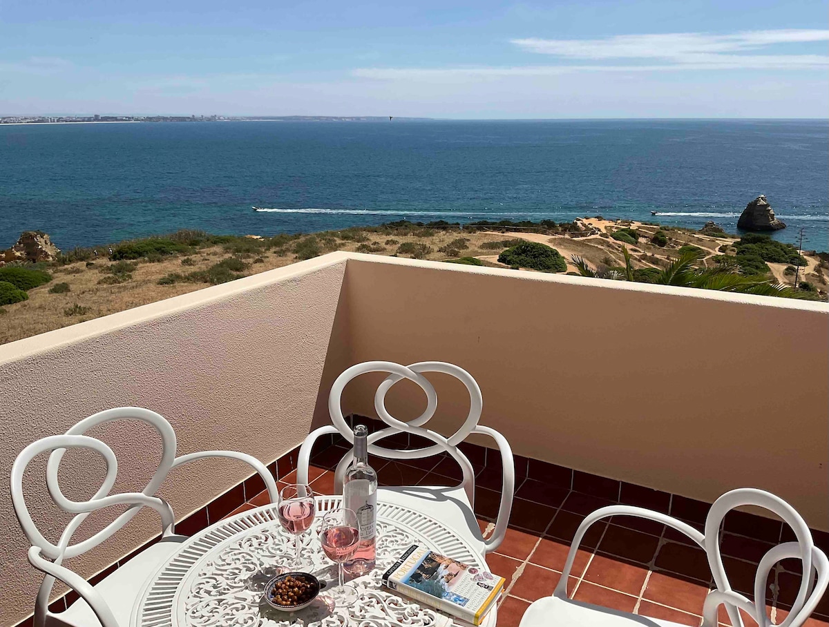 An outdoor patio is featured, equipped with a round table and four white chairs. Two glasses of pink drink are placed on the table alongside a bottle and a small dish. The backdrop showcases a panoramic view of the Atlantic Ocean and distant coastal scenery.