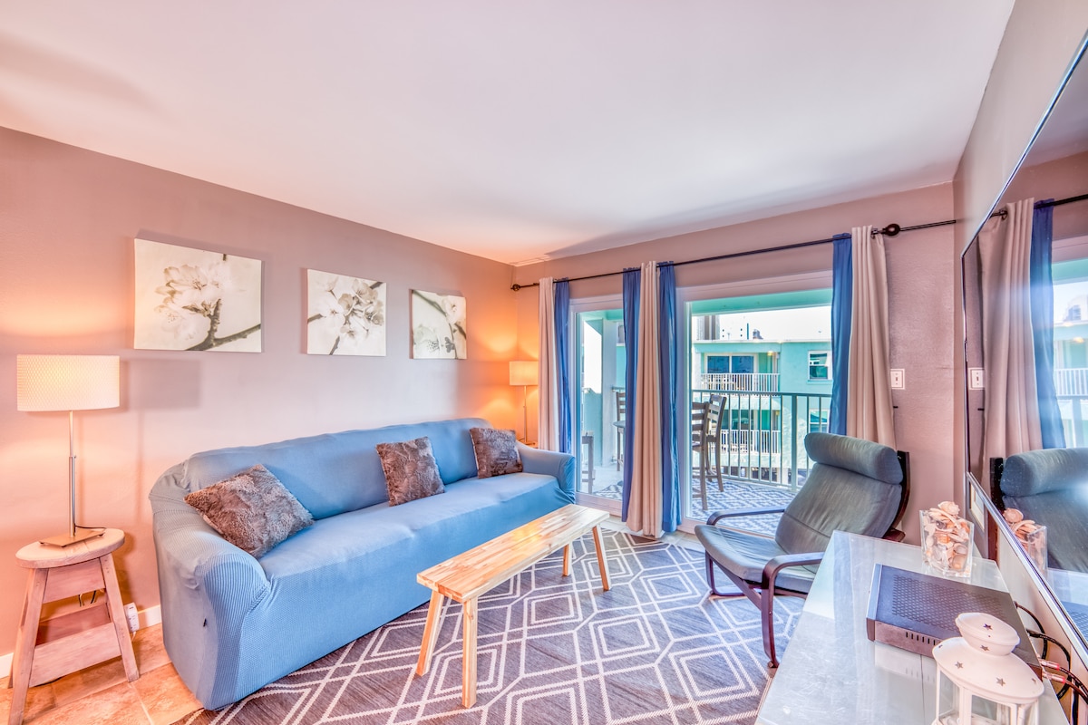 A cozy living area is furnished with a blue sofa and a coffee table, complemented by decorative artwork on the walls. Natural light flows through sliding glass doors, leading to a balcony area. A spacious rug adds warmth to the room, which includes a black chair and a TV console.