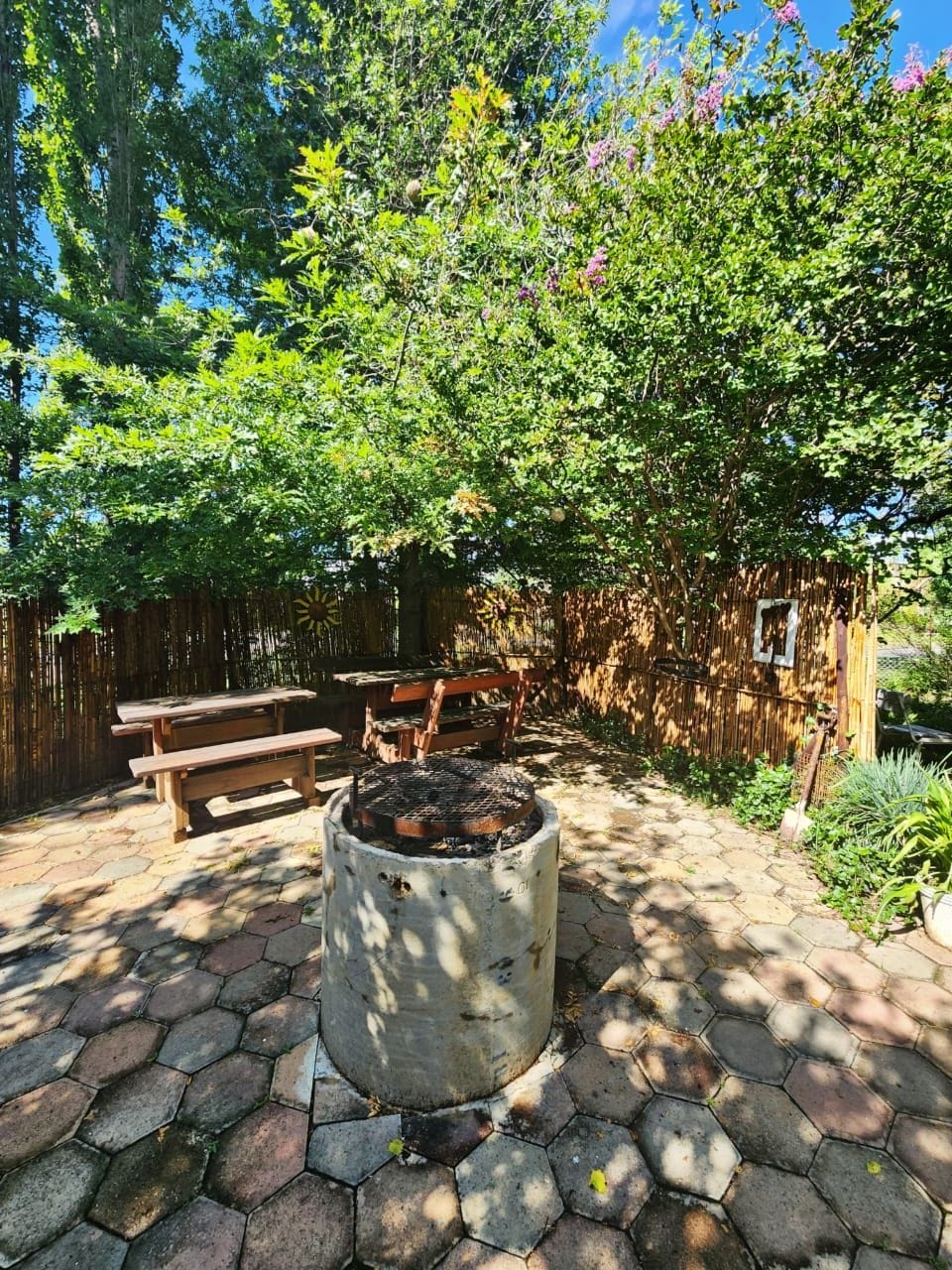 An outdoor space is featured with stone pavers and surrounded by lush greenery. A round fire pit is positioned at the center, with two wooden picnic tables nearby, offering seating for guests. Natural light filters through the trees, creating a serene atmosphere.