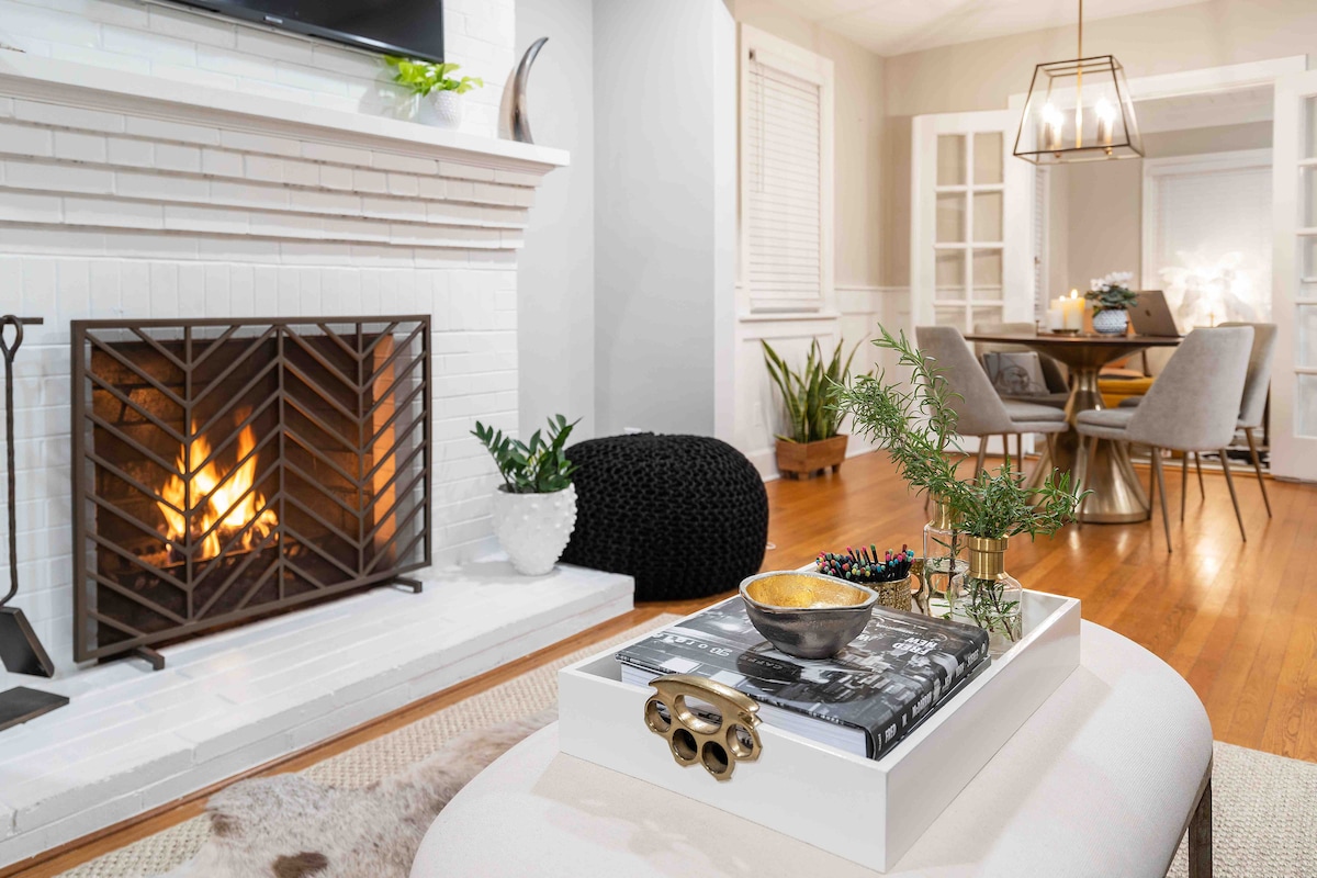 A wood-burning fireplace is framed by a white mantel, with a decorative tray placed on a light-colored ottoman. A dining area is visible in the background, featuring a round table and upholstered chairs. Soft lighting enhances the inviting ambiance of the space.