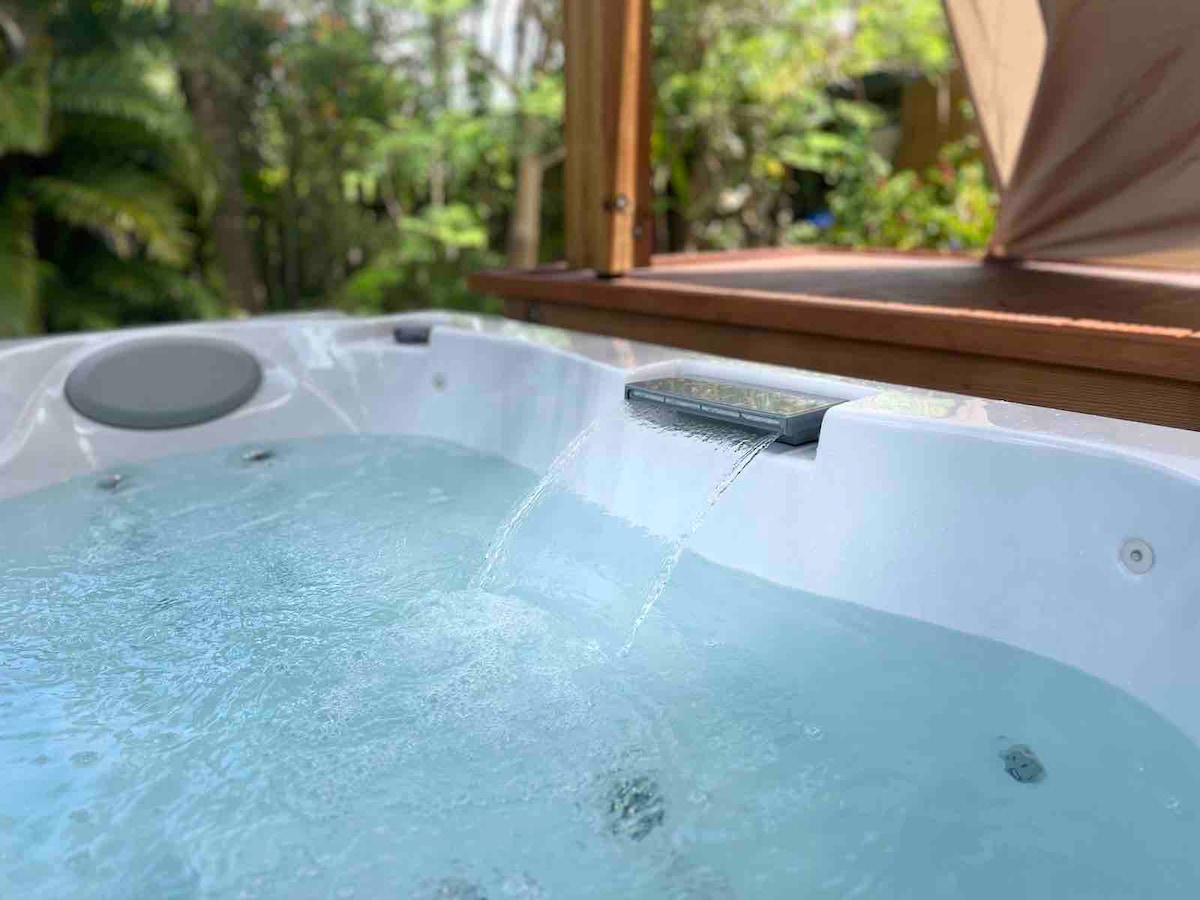 A serene jacuzzi is set in a covered terrace area, featuring a gentle waterfall that creates a soothing ambiance. Lush greenery is visible in the background, enhancing the natural surroundings of the space.