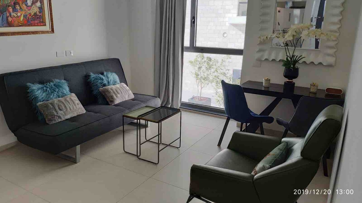 A modern living area is presented with a grey sofa adorned with decorative pillows, a glass coffee table, and a small dining table with blue chairs. Natural light enters through a large window, with greenery visible outside. The walls are kept neutral, enhancing the space's openness.