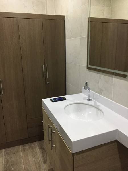 A modern bathroom features a sleek, white countertop with an integrated sink. Storage cabinets in warm wood tones are contrasted by light-colored tiles on the walls. A mirror hangs above the sink, reflecting the contemporary design. An electronic device is placed near the sink.