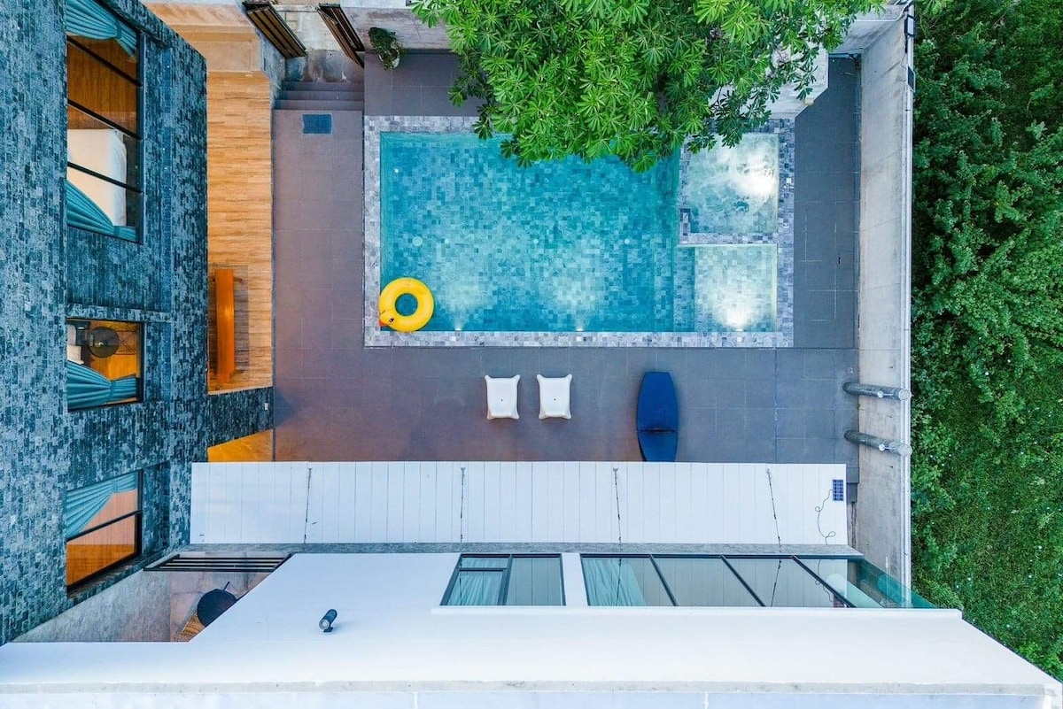 An aerial view captures a serene pool area, featuring a turquoise pool surrounded by a stone deck. Two light-colored lounge chairs are positioned nearby, accompanied by a bright yellow float. Lush green trees frame the space, highlighting the villa's tranquil ambiance.