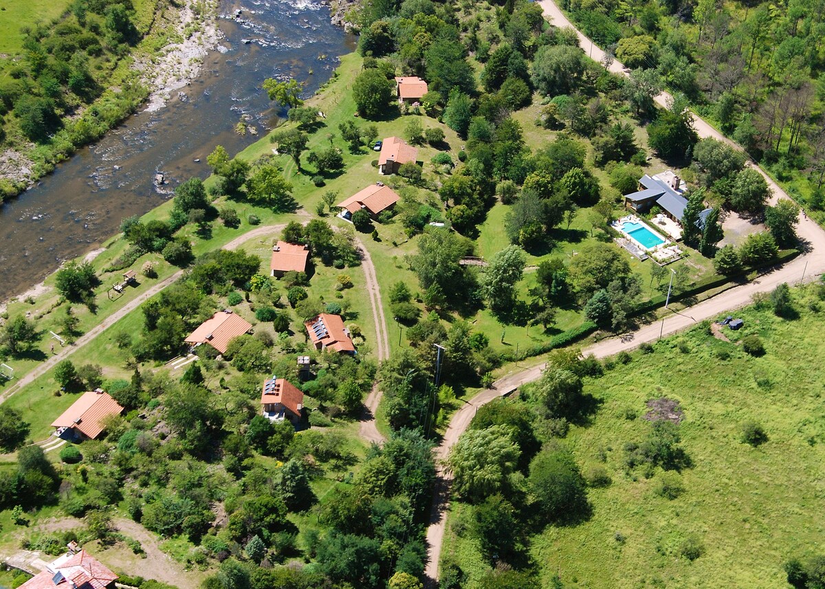 An aerial view showcases a lush green landscape with several buildings nestled among trees. A winding path is visible, leading to a pool area, while a river flows along one side of the property, offering a serene natural setting.