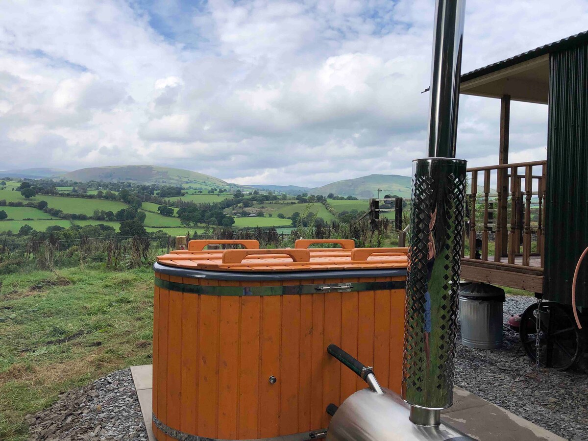 An outdoor hot tub with a wooden exterior is positioned on a stone patio. Rolling green hills are visible in the background under a partly cloudy sky, providing a serene landscape. A small wood-burning stove is nearby, complementing the tranquil atmosphere.