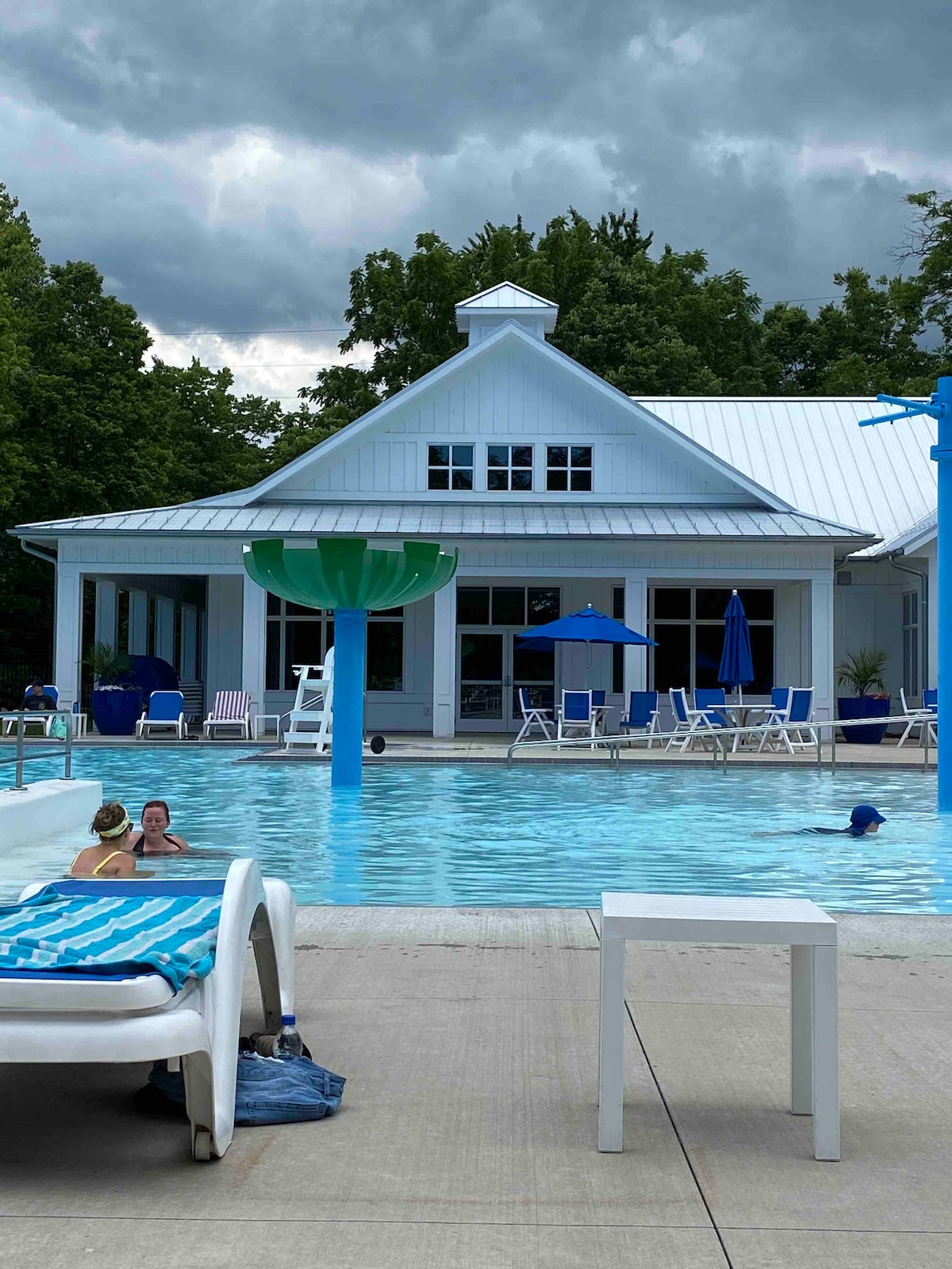 A spacious outdoor pool area is shown, with neatly arranged lounge chairs and umbrellas surrounding the pool. The backdrop features a contemporary building with large windows, and a green water slide is positioned near the pool. Dark clouds are visible in the sky.