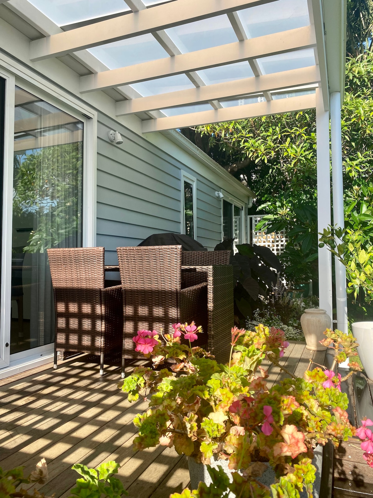 A shaded outdoor seating area is featured, complete with a wicker dining set. Lush greenery surrounds the space, with flowering plants in the foreground. Natural light filters through the overhead structure, creating a pattern on the wooden deck.