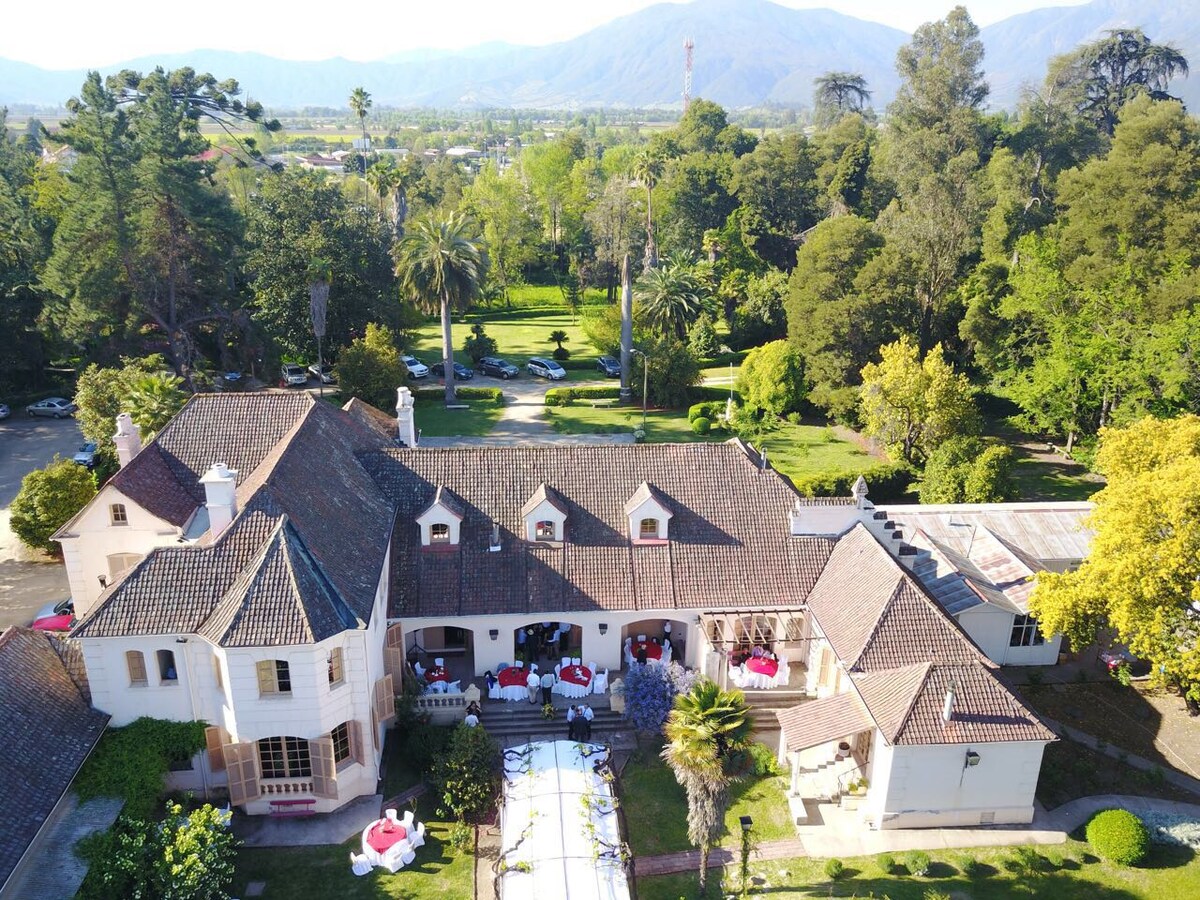 An aerial view captures the Casa Patronal, showcasing its elegant architecture and surrounding lush gardens. The expansive property features vibrant green trees and landscaped areas, with a clear view of outdoor seating arrangements placed under the shade of trees, set against a backdrop of mountains.