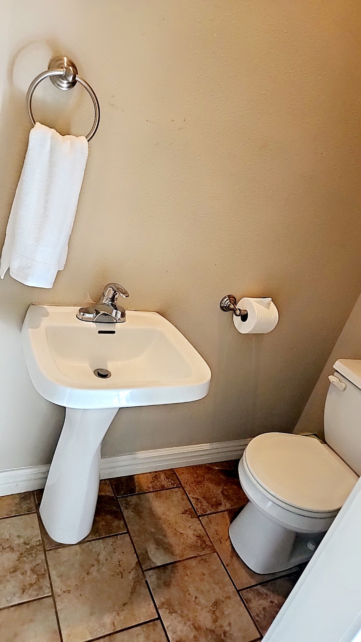 A compact bathroom is shown, featuring a modern pedestal sink with a single-lever faucet and a roll of toilet paper on a holder. A towel hangs on a ring nearby. The floor is tiled, and the walls have a neutral finish.
