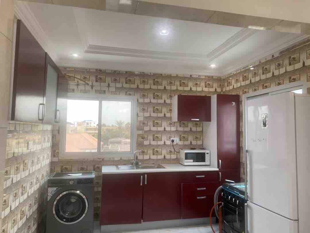 The kitchen features modern cabinetry in deep red tones, complemented by a white countertop. A large window allows for natural light, while a washing machine, refrigerator, microwave, and stove are arranged for convenient use.