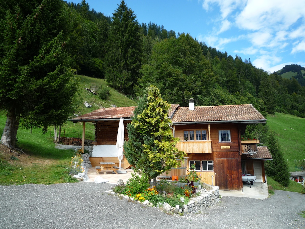 A charming chalet is nestled among green trees in a mountainous landscape. The exterior features a wooden facade and a stone path leading to a covered terrace. Colorful flowers border the gravel area, enhancing the natural setting.