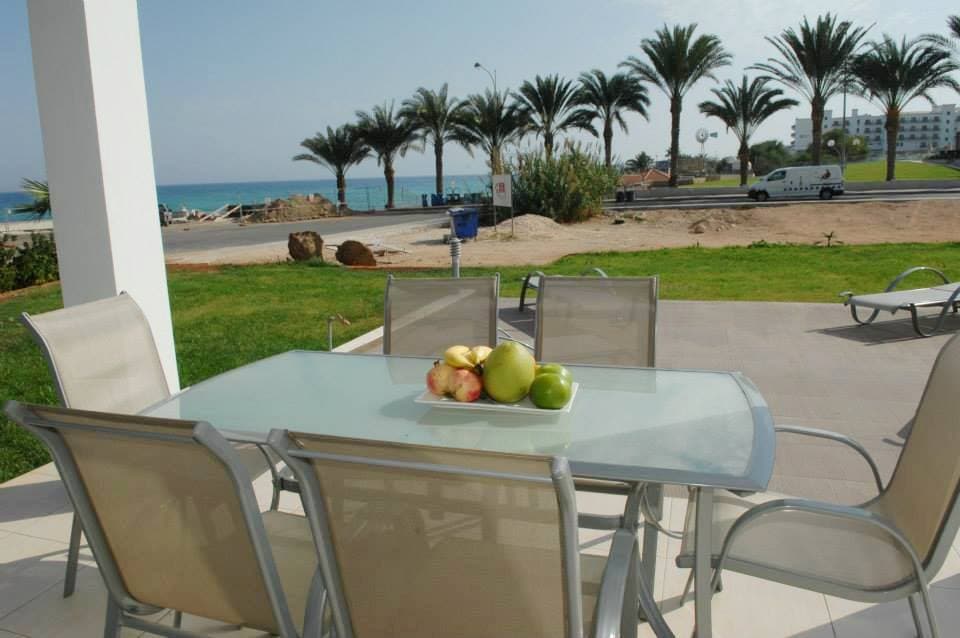 A dining table is positioned outdoors, surrounded by six metal chairs. A bowl of apples and pears sits at the center, with a view of palm trees and the sea in the background, enhancing the outdoor dining experience.