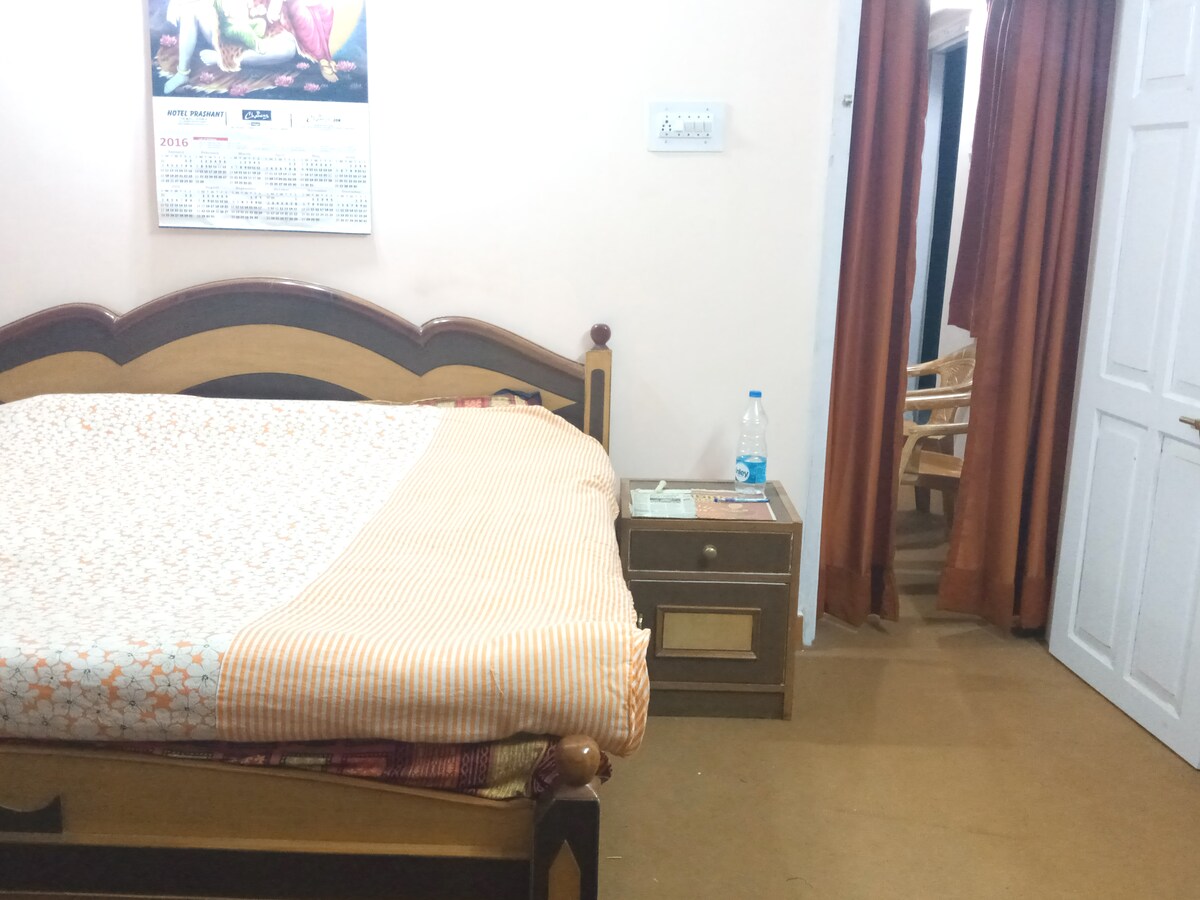A well-furnished bedroom features a spacious bed adorned with a patterned blanket. A nightstand with a water bottle rests beside the bed. In the background, there are wooden chairs and curtains that add to the room's cozy atmosphere.
