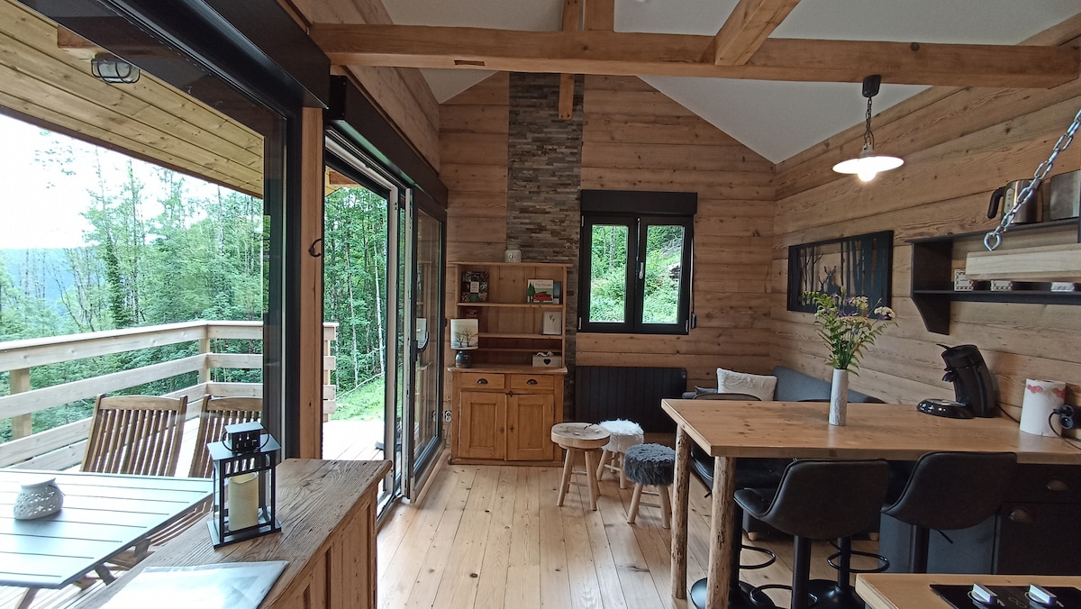 The living area features wooden walls and beams, creating a natural ambiance. Large glass doors allow for views of the surrounding greenery. A small table with high chairs is positioned next to a wooden sideboard. A cozy stool and decorative lantern enhance the inviting environment.