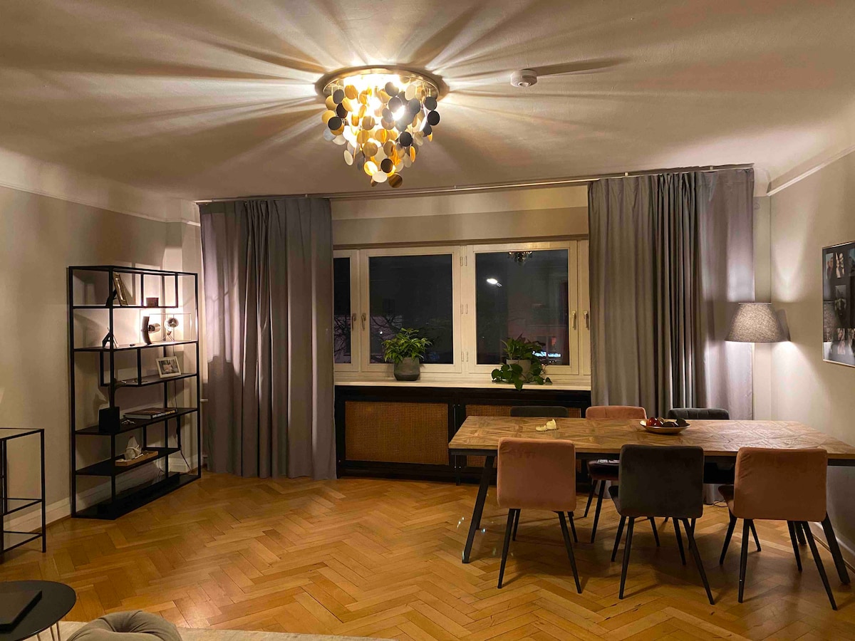 A modern living space is illuminated by a striking chandelier, casting a warm glow. A wooden dining table with several chairs is centrally positioned beneath large windows, adorned with plants. Soft curtains frame the windows, while decor and shelving units offer additional storage and visual interest.