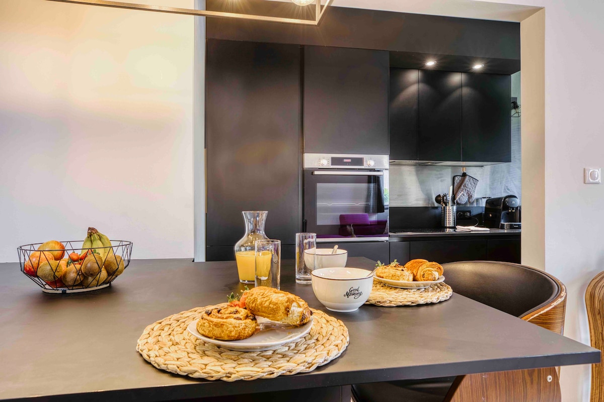 A modern kitchen space is depicted, featuring a stylish black cabinetry setup. A dining table is set with two plates holding pastries and a bowl, accompanied by a glass of orange juice and a carafe. A fruit bowl sits nearby, adding a touch of freshness.