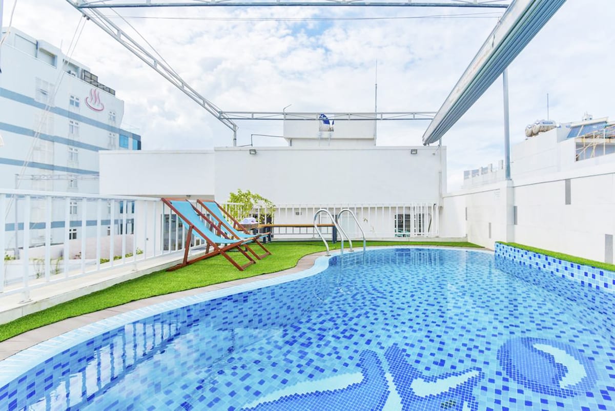 A rooftop swimming pool features blue mosaic tiles reflecting natural light. Sun loungers are arranged along the poolside on a grassy area. Nearby, large windows provide a view of the surrounding city, enhancing the open and airy atmosphere.
