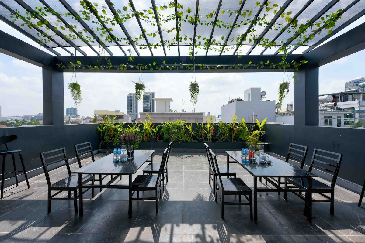 A spacious rooftop area is defined by a canopy of greenery and sunlight. Several black tables and chairs are arranged neatly, surrounded by vibrant plants and glass bottles of water. The open space offers views of the city skyline, creating an inviting outdoor setting.