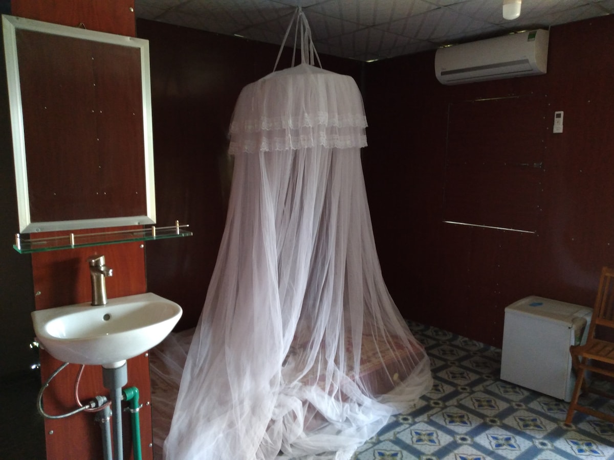 A spacious room features a large mosquito net draped over a bed, enhancing comfort. A washbasin is positioned to the left, while a small refrigerator sits discreetly on the floor. A wooden chair adds seating, and an air conditioning unit is visible on the wall.