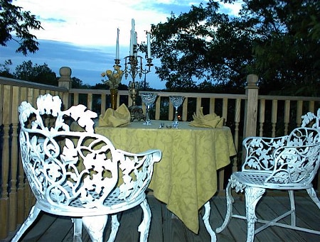 An outdoor dining area features a round table covered with a yellow tablecloth, complemented by two ornate white chairs. Elegant glassware and a candle candelabrum adorn the table, surrounded by lush greenery under an evening sky.